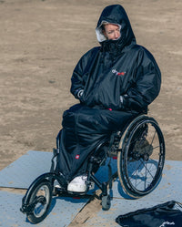 Woman sat on wheelchair on a beach, wearing dryrobe® Adapt with the hood up