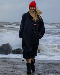 Woman walking on beach with sea behind her, wearing dryrobe Advance Long Sleeve changing robe in Black Red and Red dryrobe Beanie