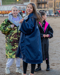 Three girls laughing at the camera stood on a beach, wearing Kids dryrobe Advance change robes