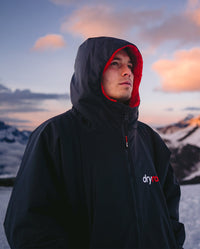 *MALE* Man with stood in front of snowy mountains, wearing dryrobe Advance Long Sleeve changing robe in Black Red with hood up