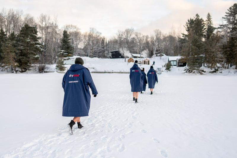 Back view of four people wearing custom Unbounded and Dryrobe® co-branded changing robes, walking through deep snow towards a winter campsite and cabins in the Canadian woods.