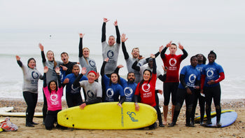 A group of Wave Project volunteers and participants taking a group photo on the beach with a yellow foam surfboard lying horizontally on the floor