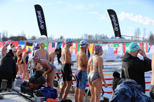 Competitors in colourful swimwear and caps preparing for their heat on the ice-covered pool deck, lined with international flags and Dryrobe® banners.