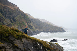 A woman in a grey and orange Dryrobe stood holding a surfboard by a cliff