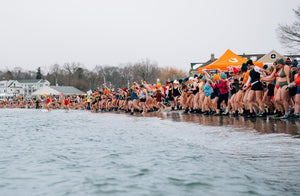 A wide shot of a massive crowd of women in swimwear and winter accessories lined up on a snowy beach, holding hands and preparing to charge into the cold water.