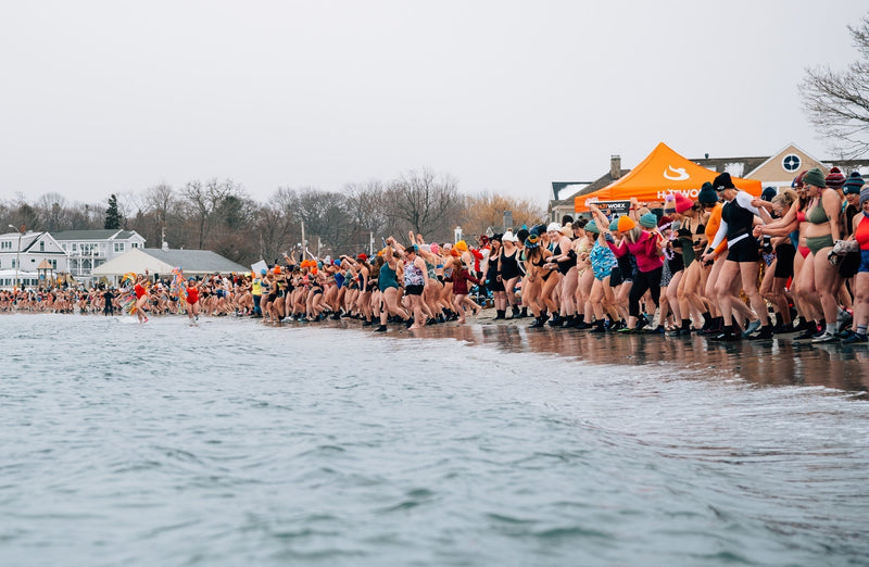 A wide shot of a massive crowd of women in swimwear and winter accessories lined up on a snowy beach, holding hands and preparing to charge into the cold water.
