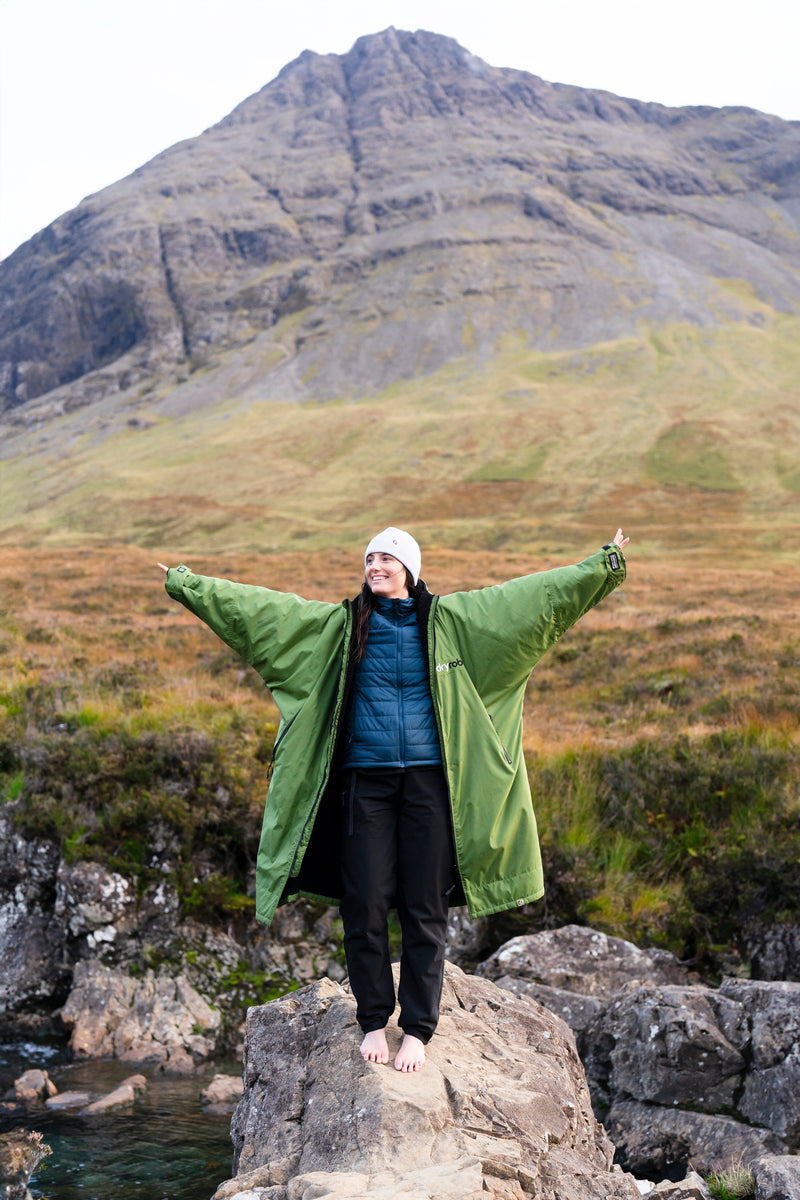 A woman stood on a rock near a fairy pool in Scotland wearing a green Dryrobe and her hands in the air
