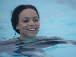 A serene, medium shot of Cat White smiling peacefully while treading water in a pool. She is wearing a black swimsuit, with the calm, light blue water reflecting on her face.