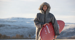 Louis Thomas-Hudson in a grey Dryrobe holding a red surfboard with a snowy beach scene in the background