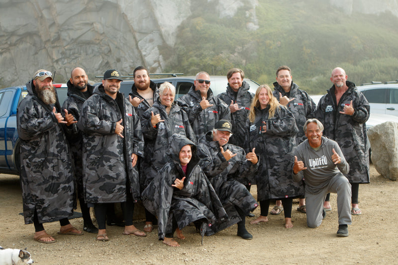 A group of people posing and smiling towards the camera on the beach wearing matching Black Camo Black dryrobes