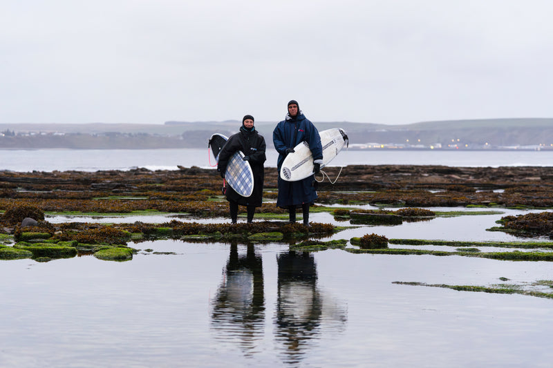 A male and female surfer stood on rocks holding surfboards and wearing dryrobes 