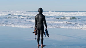 A surfer in a full black wetsuit and hood stands on a sunlit beach, looking out at the ocean waves while holding a camera housing and fins.