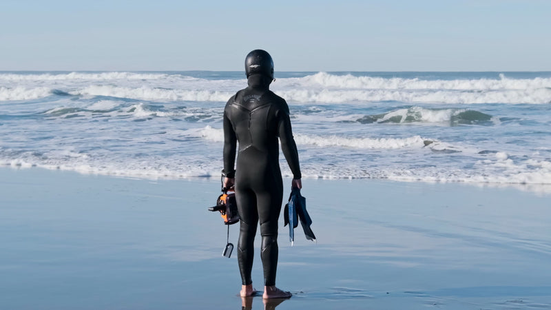A surfer in a full black wetsuit and hood stands on a sunlit beach, looking out at the ocean waves while holding a camera housing and fins.