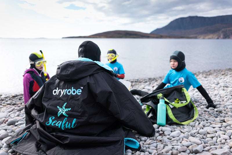 A group of children sat on a pebble beach wearing snorkelling gear with a Seaful branded Dryrobe wrapped around one of them