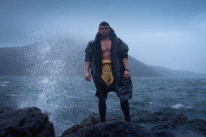 Ross Edgley stood in a black camo dryrobe and a wetsuit on a rock as a splash from a wave hits it 
