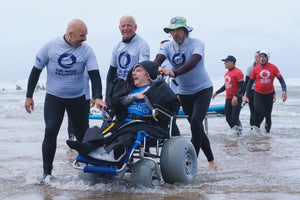 An adaptive surfer in a beach wheelchair laughing while being assisted into the ocean by The Wave Project volunteers