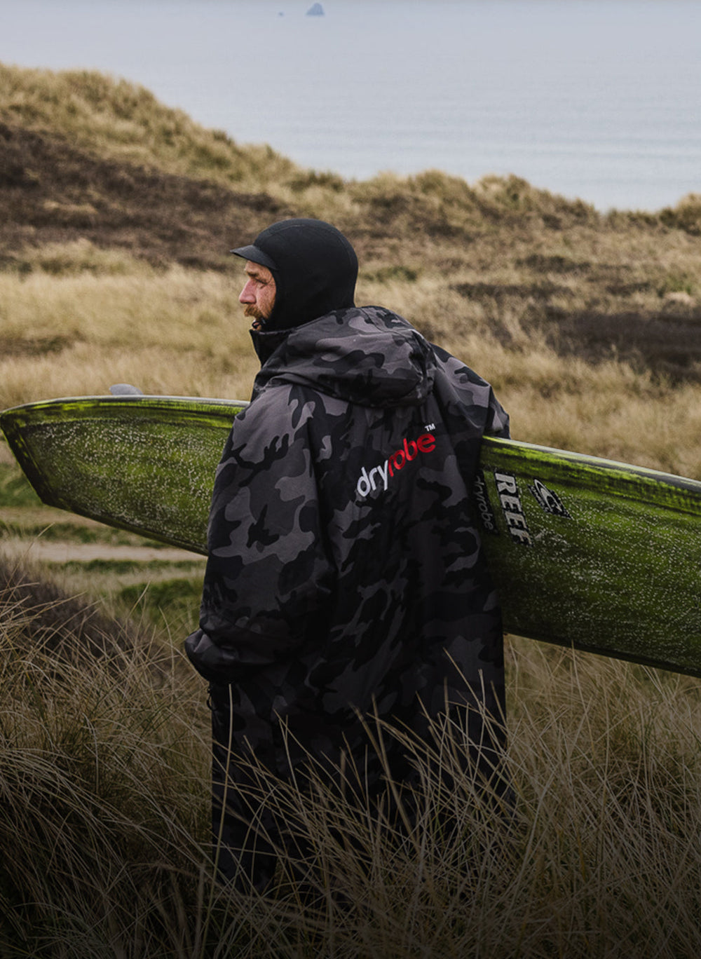 Adult male surfer wearing black camo Dryrobe® long sleeve holding surfboard walking to beach 