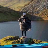 Person wearing a Dryrobe® changing robe standing on a rock by a lake with mountains in the background