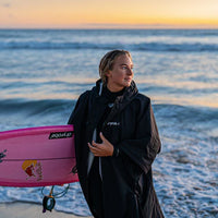 Person wearing a Dryrobe change robe standing with a surf board by the sea