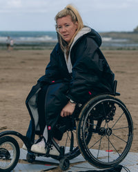 Woman sitting on wheelchair on a beach, wearing dryrobe® Adapt