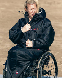 Woman sat on wheelchair on a beach, wearing dryrobe® Adapt with the hood down