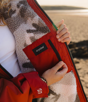 Close-up of a person at a beach wearing a red Dryrobe Changing robe, with a cream sherpa lining and a black brand patch on the chest pocket.