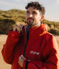 *MALE* A man wearing a bright red Dryre waterproof Changing robe, stands on a sandy beach with dunes in the background. The white 'dryrobe' logo is visible on the left chest of the Changing robe,.