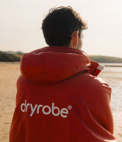 *MALE* A man viewed from behind wearing a red hooded Dryrobe Changing robe, on a sandy beach with water in the background.