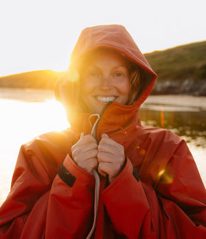 A smiling woman wearing a bright red hooded waterproof Changing robe, stands outdoors by the water at sunset, holding the front zipper closed with both hands.