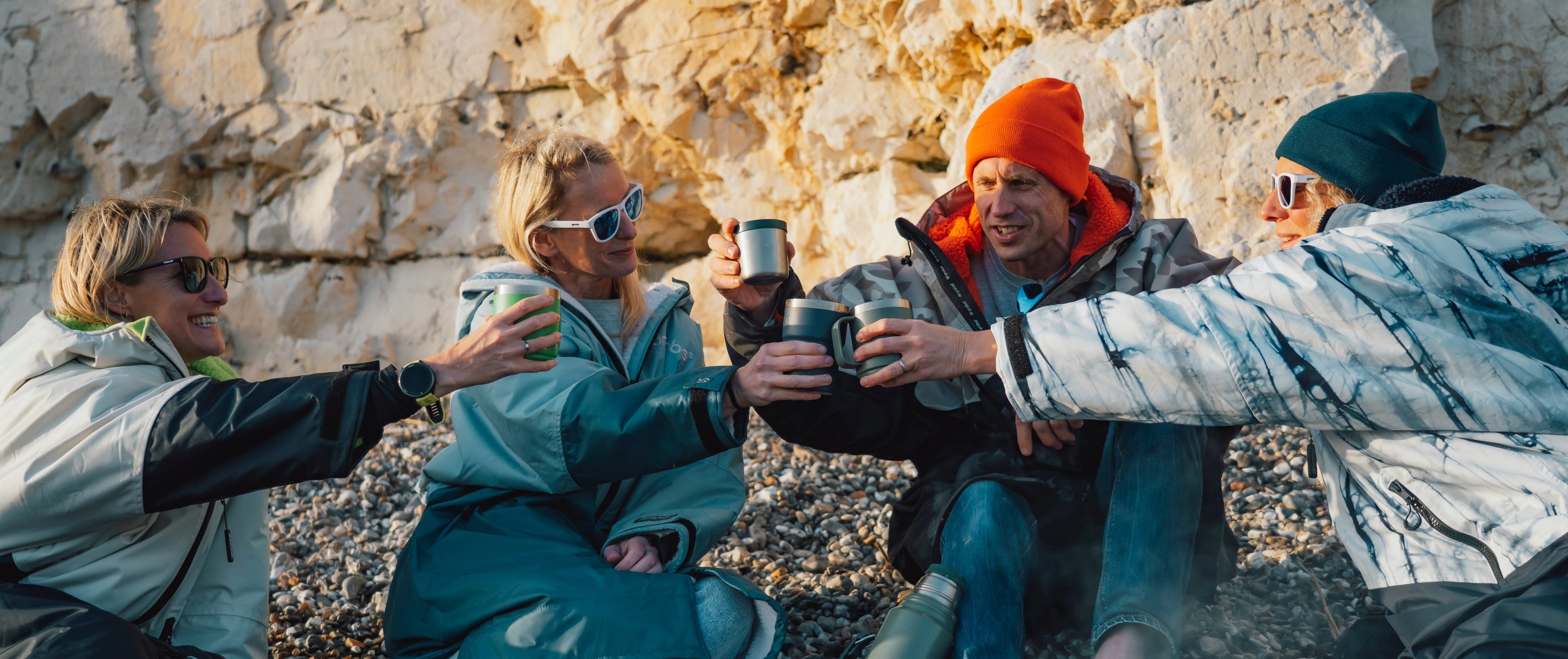 Four people in dryrobes sitting together, holding drinks against a rocky background.