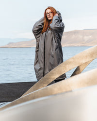 Woman stood in front of lake wearing dryrobe Advance Long Sleeve Alpine Range change robe in Alpine Graphite, pulling hood up