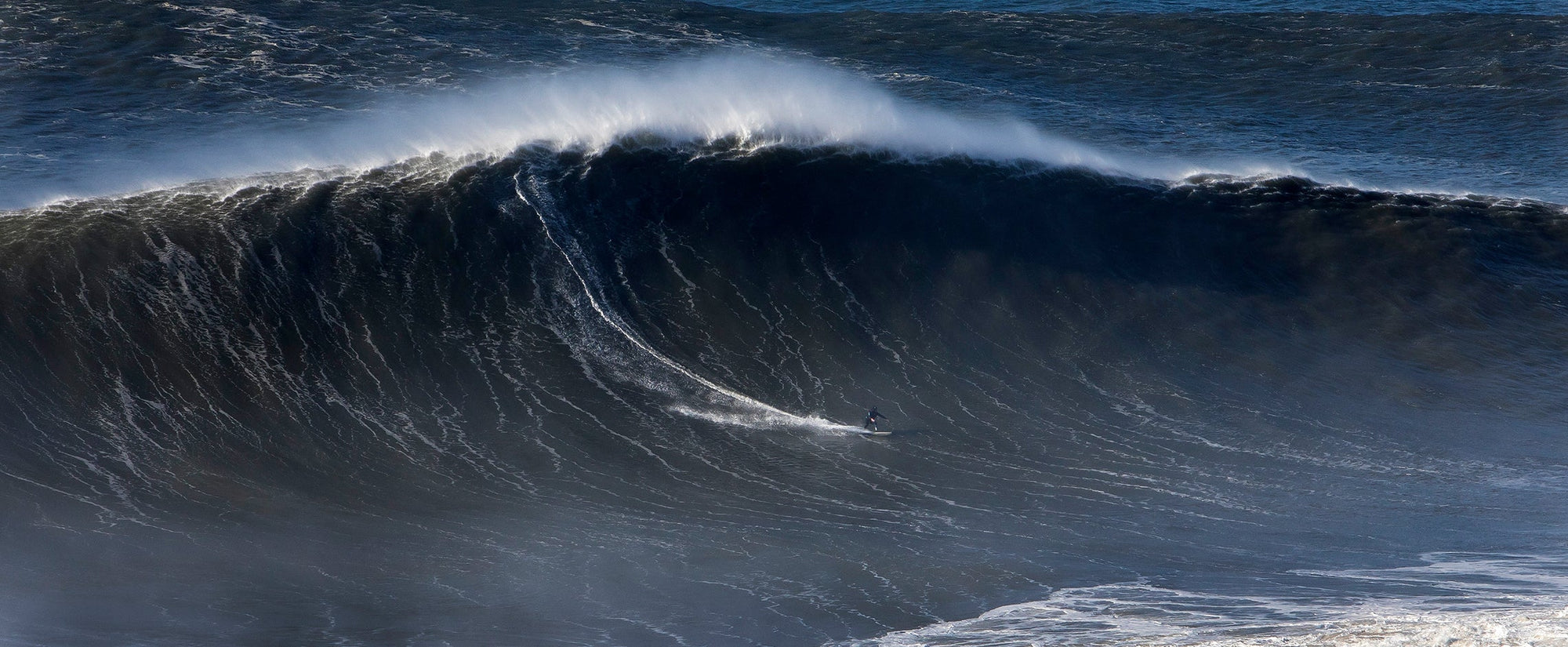 Large wave crashing in the ocean with a dark blue color. Big wave surfing by Andrew Cotton.