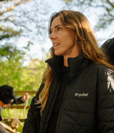 A woman outdoors wearing a black Dryrobe puffer jacket, shown in a close-up profile view with sunlight filtering through trees.
