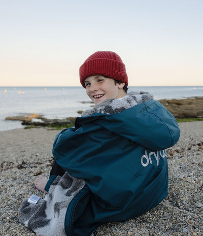 *MALE* A smiling boy sitting on a pebble beach, wrapped in a teal hooded Dryrobe with a gray and white faux-fur trim and a red knit beanie.