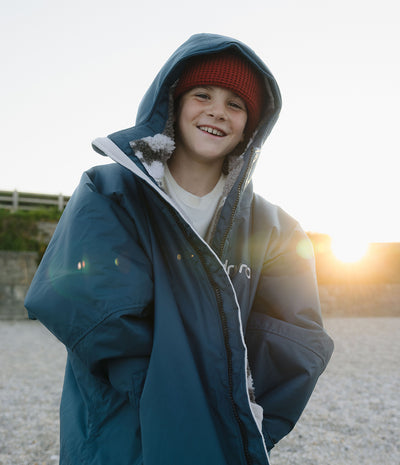 *MALE* A smiling young boy wearing a navy blue hooded Dryrobe with white fleece lining and a red knit beanie, standing outdoors in bright sunset light.