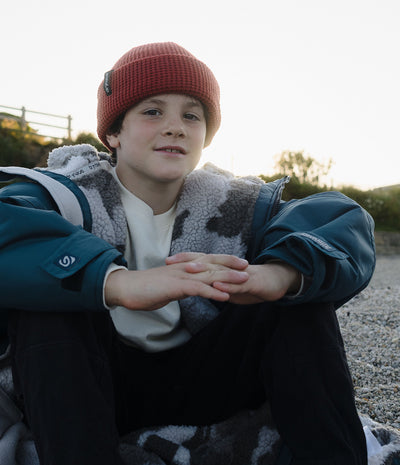 *MALE* A young boy sitting outdoors on a gravel surface, wearing a rust-red knit beanie, a teal and Dryrobe over a white shirt, and black pants.
