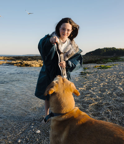 A woman wearing a dark teal hooded Dryrobe with a shearling-style collar stands on a pebble beach holding a leash attached to a golden retriever dog, with water, rocks, and a clear blue sky in the background.