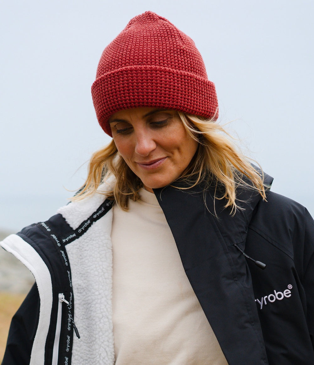 A woman wearing a rust-red knit beanie, a cream turtleneck, and a black and white Dryrobe jacket, photographed outdoors against a soft, overcast sky.