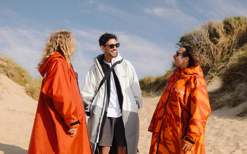Three people standing on a sandy dune wearing oversized waterproof poncho-style jackets in bright orange and white tones under a blue sky.