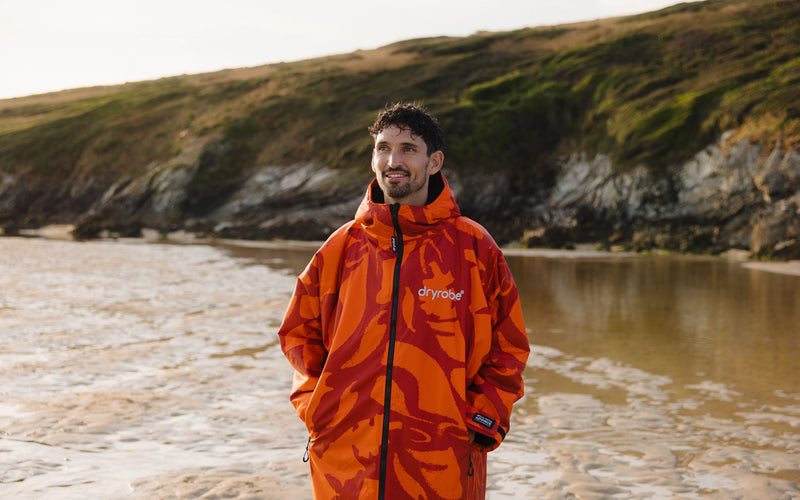 A man wearing a bright orange and red patterned Dryrobe hooded jacket stands on a sandy beach with rocky, grassy cliffs in the background.
