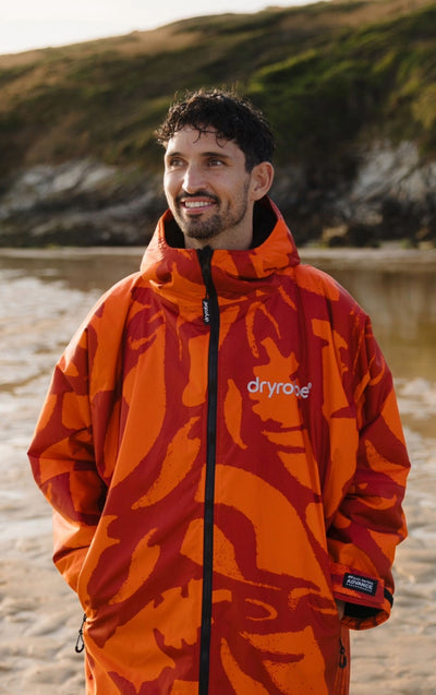A man wearing a bright orange and red abstract patterned dryrope waterproof jacket, standing on a sandy beach with rocky cliffs and water in the background.
