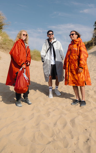 Three models standing on a sandy dune wearing long rain ponchos in orange and gray, with a blue sky in the background.
