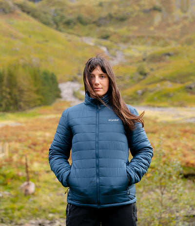 A woman wearing a blue puffer jacket stands in a grassy mountain landscape. The jacket has a small white Dryrobe logo on the left chest.