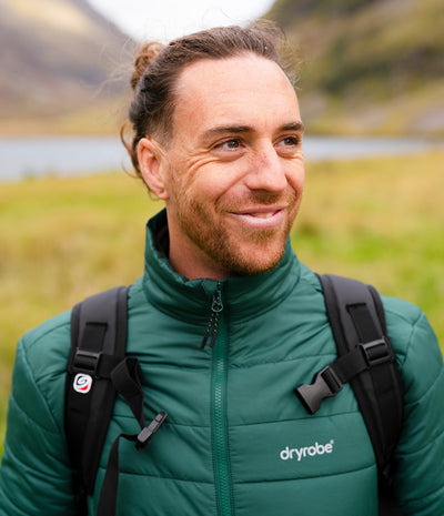 A man outdoors wearing a green Dryrobe puffer jacket and a black backpack, with a blurred natural landscape in the background.
