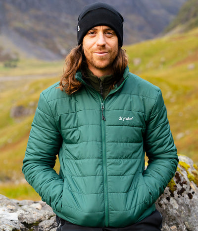 A man wearing a green dryrobe puffer jacket and a black beanie stands outdoors against a blurred mountain and grassy landscape background.