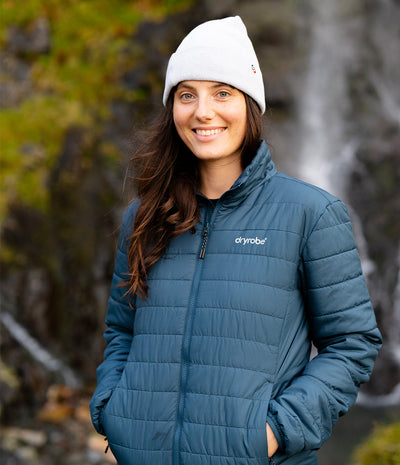 A woman wearing a slate blue puffer jacket and a white beanie stands outdoors in front of a blurred waterfall and rocky background. The jacket has the 'dyrobe' brand name visible on the left chest.