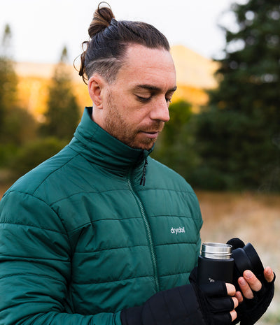 A man wearing a green Dryrobe puffer jacket and black gloves holds a stainless steel travel mug outdoors with a blurred natural background.