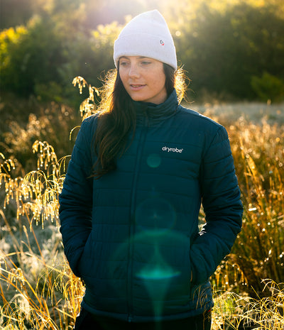 A woman wearing a white beanie and a dark teal Dryrobe puffer jacket stands in a sunlit field of dry grass.