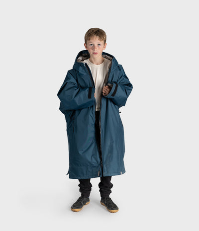*MALE* A young boy wearing a long, oversized dark blue hooded Dryrobe over a cream t-shirt, black pants, and black sneakers, standing against a light gray studio background.
