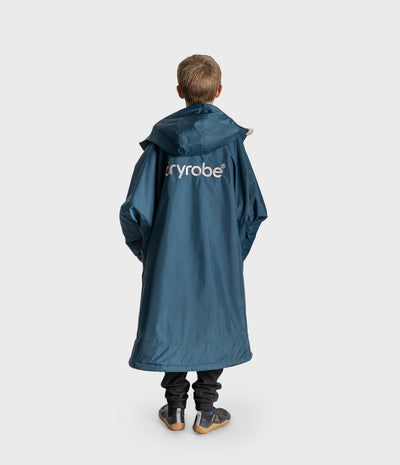 *MALE* A child viewed from behind wearing a long teal-blue hooded Dryrobe with the brand name 'Dryrobe' printed in white across the upper back, paired with dark pants and sneakers, against a light gray studio background.